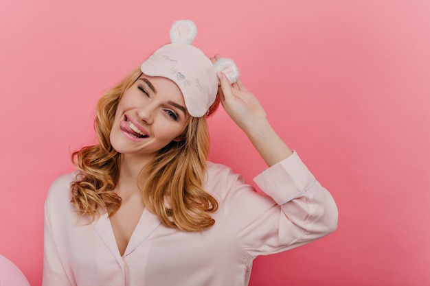 Alegre jovencita con cabello rubio brillante posando con una bonita sonrisa. Foto interior de la alegre niña positiva en máscara de dormir aislada en la pared rosa.