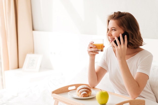 Alegre jovencita sentada en el interior con croissant beber jugo