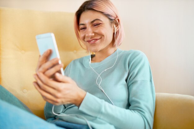 Alegre linda joven europea con piercing facial relajante en casa con teléfonos inteligentes.