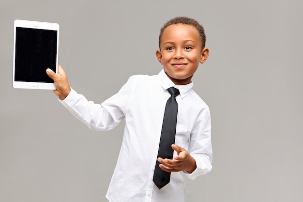 Alegre lindo colegial afroamericano en camisa y corbata sonriendo felizmente usando un dispositivo electrónico para jugar o ver dibujos animados, sosteniendo una tableta digital con pantalla en blanco con copyspace para su texto