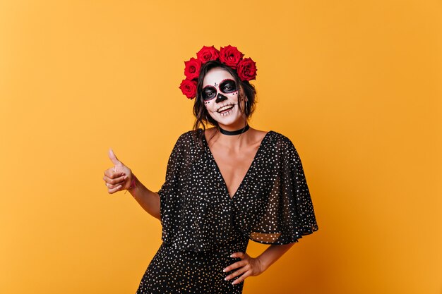 Alegre lindo mexicano con cabello oscuro levanta los pulgares. Retrato de niña con maquillaje inusual en un hermoso vestido negro.