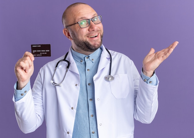 Alegre médico de mediana edad vistiendo bata médica y un estetoscopio con gafas con tarjeta de crédito que muestra la mano vacía aislada en la pared púrpura