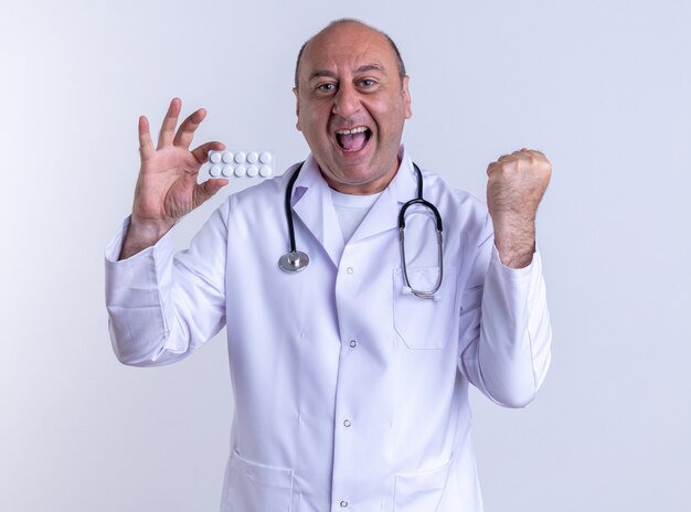 Alegre médico de mediana edad vistiendo una bata médica y un estetoscopio que muestra el paquete de tabletas mirando al frente haciendo un gesto de sí aislado en la pared blanca
