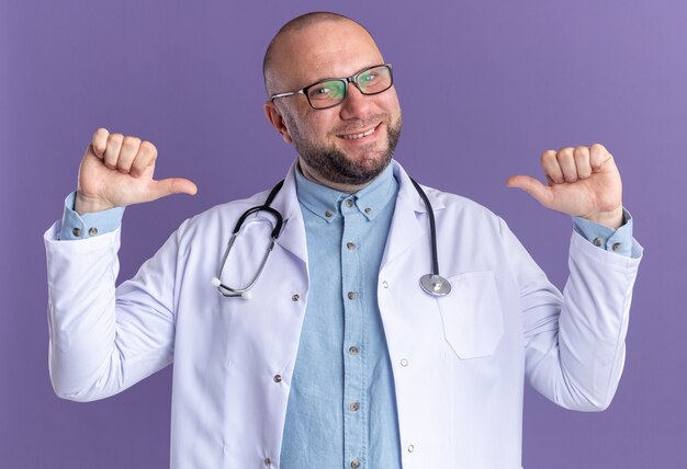 Alegre médico varón de mediana edad vistiendo bata médica y estetoscopio con gafas apuntando a sí mismo aislado en la pared púrpura