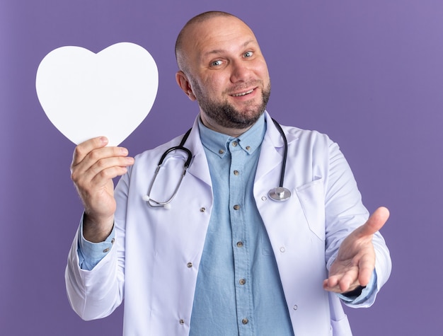 Alegre médico varón de mediana edad vistiendo bata médica y un estetoscopio sosteniendo en forma de corazón mostrando la mano vacía