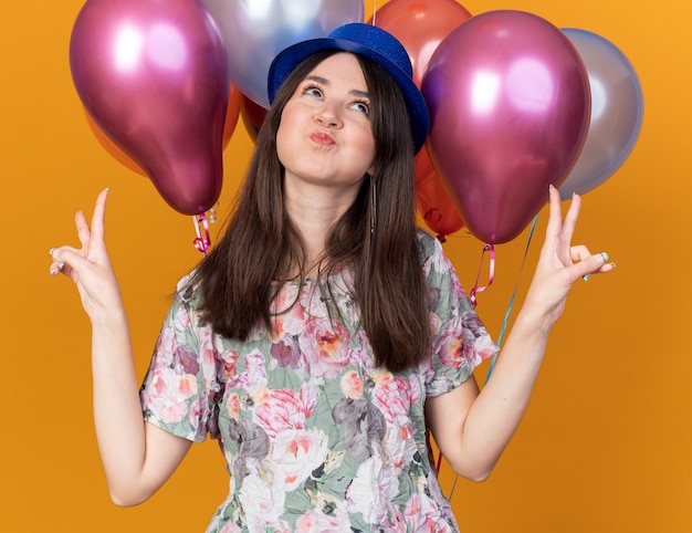 Alegre mirando hacia arriba joven hermosa chica con sombrero de fiesta de pie delante de globos mostrando gesto de paz aislado en la pared naranja