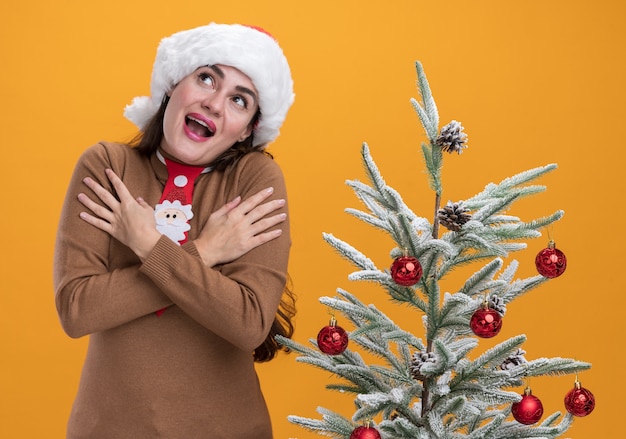 Alegre mirando hacia arriba joven hermosa niña con sombrero de navidad con corbata de pie cerca del árbol de navidad poniendo las manos en el hombro aislado en la pared naranja