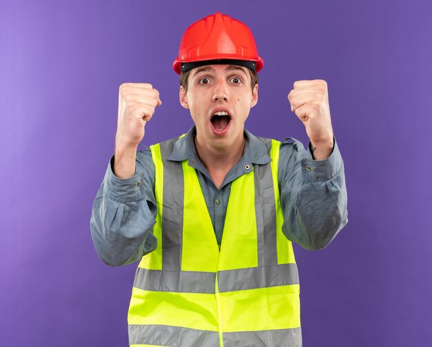 Alegre, mirando a la cámara, joven constructor hombre en uniforme mostrando sí gesto