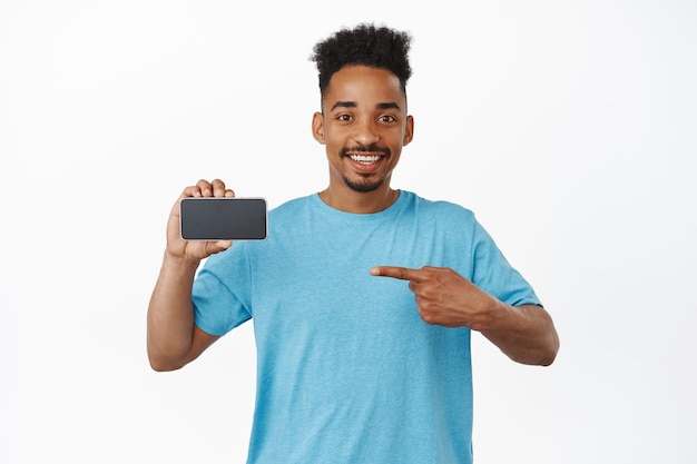 Alegre modelo afroamericano masculino señalando con el dedo la pantalla horizontal del teléfono móvil, recomendando tienda, aplicación en smartphone, interfaz de espectáculo, fondo blanco