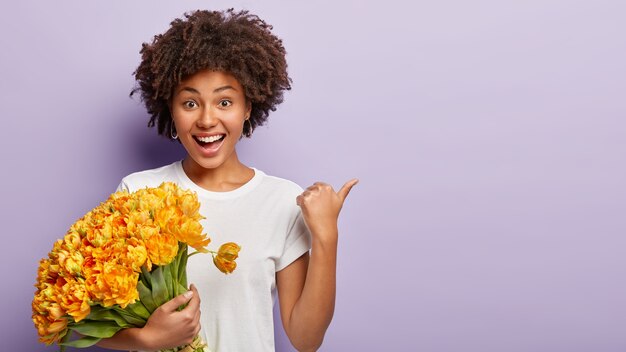 Alegre modelo femenino de piel oscura apunta a un lado en el espacio en blanco, sostiene un ramo de tulipanes naranjas de primavera