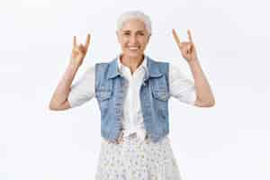 Foto gratuita alegre, moderna y elegante abuela que se divierte con sus nietos escuchando música rock and roll contando buenos viejos tiempos en conciertos de heavy metal mostrando signos de rock y sonriendo despreocupado fondo blanco