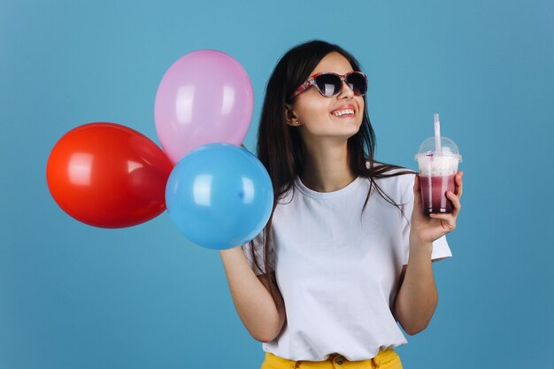 Alegre morena en gafas de sol negras se ve feliz posando con un cóctel y globos