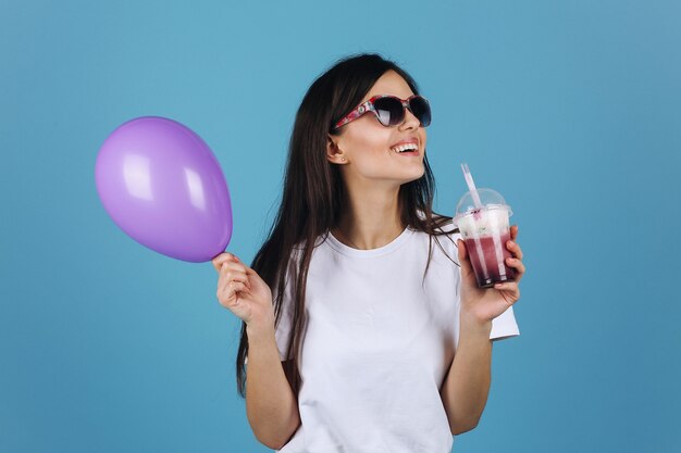 Alegre morena en gafas de sol negros se ve feliz posando con un cóctel y un globo