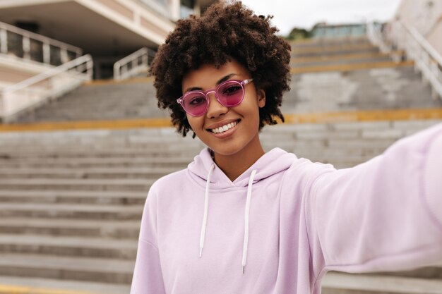 Alegre morena rizada mujer de ojos marrones con gafas de sol rosas y sudadera con capucha púrpura sonríe y toma selfie cerca de las escaleras afuera