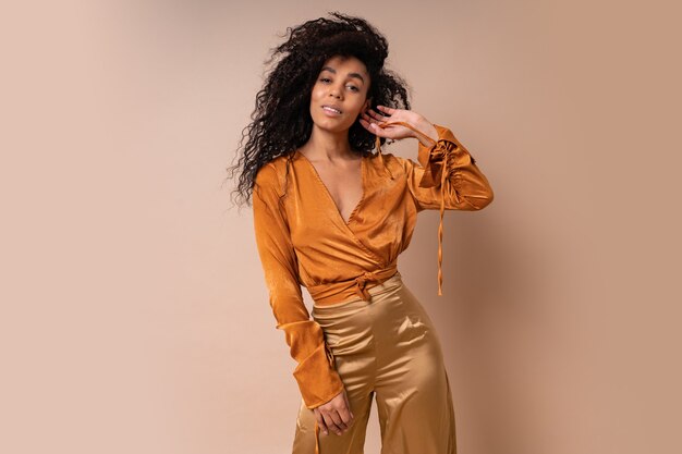 Alegre mujer africana con cabellos rizados perfectos en blusa naranja casual y pantalón dorado posando en la pared beige.