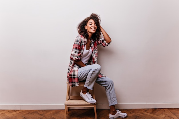 Foto gratuita alegre mujer africana sentada en la luz con una sonrisa. chica muy bien vestida disfrutando de disparos en interiores.