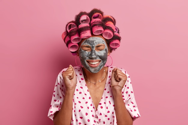 Foto gratuita alegre mujer afroamericana aprieta los puños, espera el efecto agradable de la máscara de arcilla, cierra los ojos y sonríe ampliamente, se aplica rulos, se viste informalmente, posa