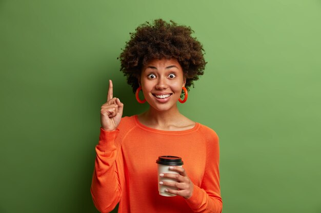 Alegre mujer afroamericana con cabello rizado, bebe café para llevar, señala con el dedo índice arriba, satisfecha de visitar la acogedora cafetería, tiene una gran sonrisa, recomienda algo, posa sobre una pared verde