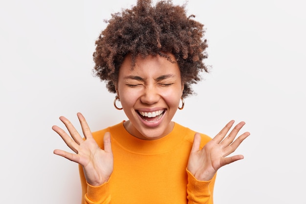 Alegre mujer afroamericana expresa felicidad, sonríe ampliamente, mantiene los ojos cerrados, levanta las palmas, tiene un estado de ánimo optimista, envía vibraciones positivas vestidas de manera informal, aisladas sobre una pared blanca. Concepto de emociones