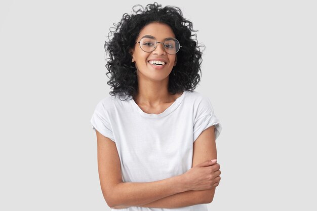 Alegre mujer afroamericana mantiene las manos cruzadas, se ríe de un buen chiste, viste ropa casual y gafas redondas, aisladas. Feliz mujer joven con piel oscura posa interior