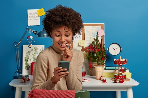 Alegre mujer afroamericana mira con alegría la pantalla del teléfono inteligente, envía un mensaje a su compañero de grupo, discute la preparación del examen en el chat en línea