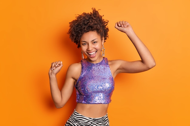 Alegre mujer afroamericana de moda con cabello rizado baila despreocupada vestida con la parte superior brillante púrpura levanta los brazos aislados sobre la pared naranja vívida