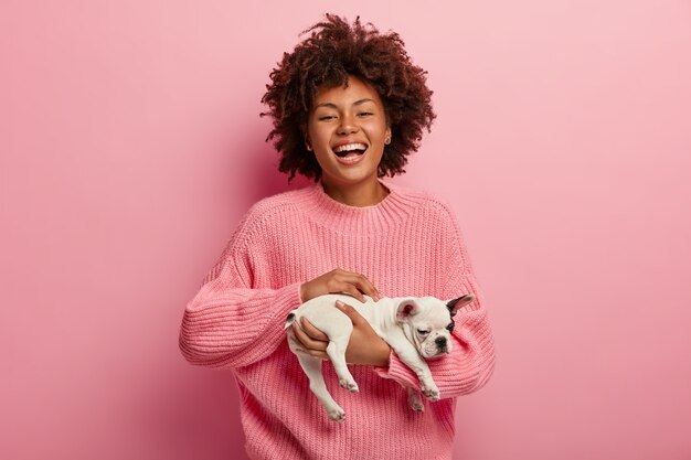 Alegre mujer afroamericana con peinado rizado, abraza a un cachorrito, muy animada, viste un jersey rosa de gran tamaño en un tono con la pared. Concepto de cuidado de mascotas y animales. Encantador miembro de la familia