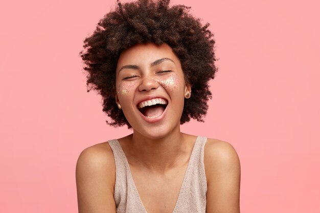 Alegre mujer afroamericana con piel oscura, se ríe alegremente, abre la boca ampliamente, tiene destellos en las mejillas, cierra los ojos, tiene el pelo rizado, aislado sobre una pared rosada. Concepto de personas y felicidad