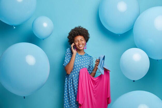 Alegre mujer afroamericana se prepara para charlas de fiesta con un amigo a través de un teléfono inteligente elige el atuendo para usar sostiene el vestido en perchas y zapatos de tacón alto