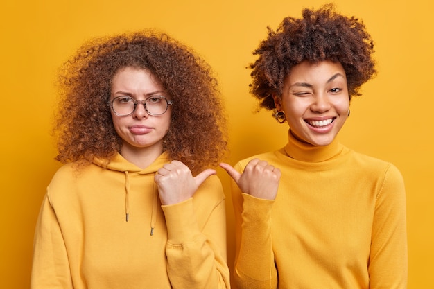 Alegre mujer afroamericana y su triste hermana de pelo rizado señalan con el pulgar el uno al otro expresan diferentes emociones vestidas casualmente aisladas sobre una pared amarilla. Es ella. Dos mujeres, interior