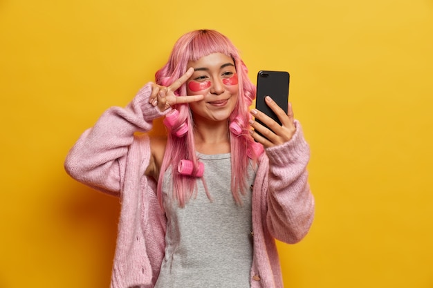 Alegre mujer asiática con cabello rosado, hace un gesto de paz sobre los ojos, se toma una selfie, se aplica parches de colágeno debajo de los ojos