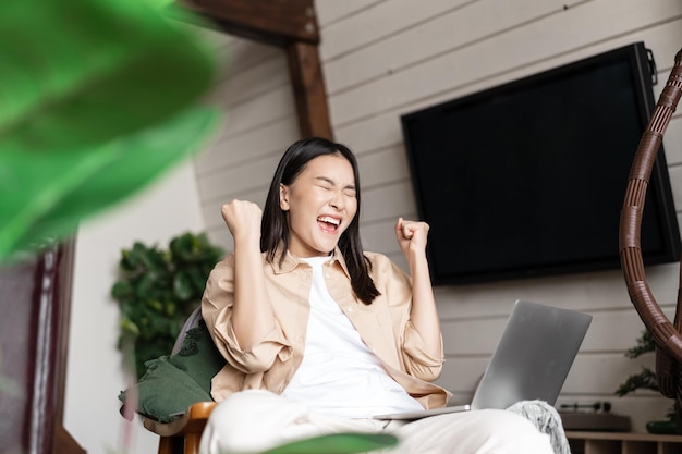 Alegre mujer asiática ganando en línea en la computadora portátil celebrando la victoria gritando y regocijándose triunfando