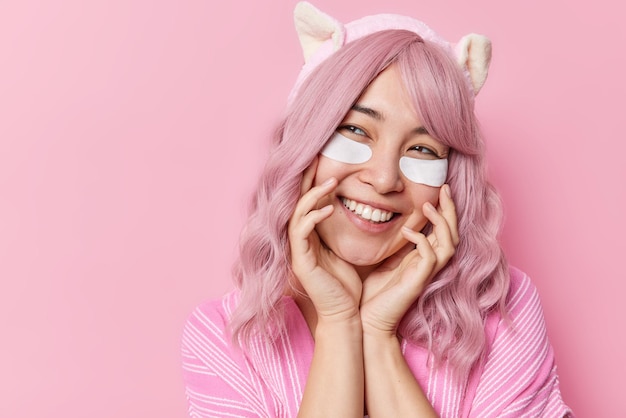 Una alegre mujer asiática de pelo rosa mantiene las manos en la cara sonriendo alegremente aplica parches de belleza blanca debajo de los ojos lleva una diadema vestida con ropa informal aislada sobre un espacio en blanco de fondo rosa