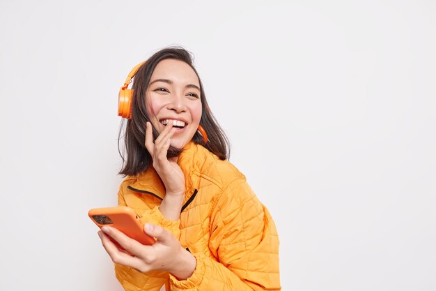 Alegre mujer asiática se ríe felizmente mira hacia otro lado en la distancia disfruta de la lista de reproducción de música favorita usa la aplicación móvil usa auriculares inalámbricos vestidos con una chaqueta naranja aislada en un espacio en blanco de la pared blanca