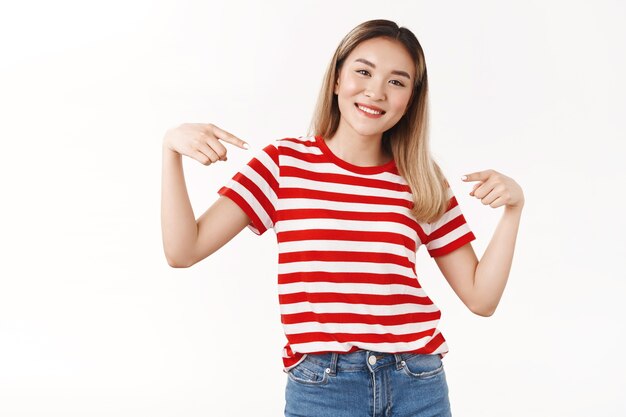 Alegre mujer asiática rubia atractiva positiva apuntando a sí misma con los dedos índices indican logros personales alardear sonriendo ampliamente fue elegida presentando con orgullo, pared blanca