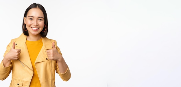 Foto gratuita alegre mujer asiática sonriendo complacida mostrando thumbs up en aprobación de pie en ropa casual sobre w