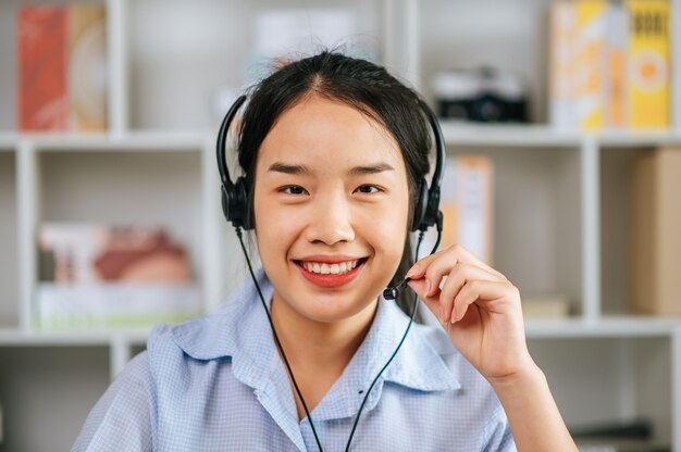 Alegre mujer asiática usa auriculares sonriendo y usa la conferencia de transmisión de videollamadas de la computadora portátil para trabajar en línea durante la cuarentena covid-19 autoaislamiento en casa, trabajar desde el concepto de hogar
