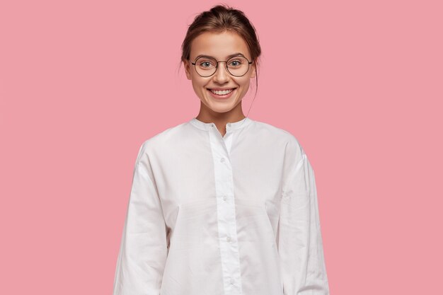 Alegre mujer de aspecto agradable en gafas, tiene expresión feliz, vestida con camisa blanca
