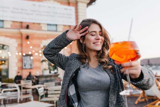 Alegre mujer blanca posando con los ojos cerrados sosteniendo una copa de cóctel de naranja