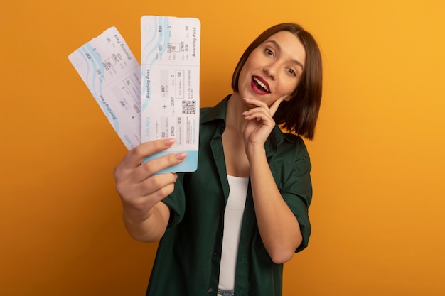 Alegre mujer bonita caucásica pone la mano en la cara y sostiene boletos de avión en naranja