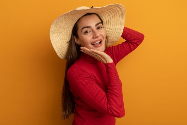 Alegre mujer bonita caucásica con sombrero de playa con la mano en la cara y mira a la cámara en naranja