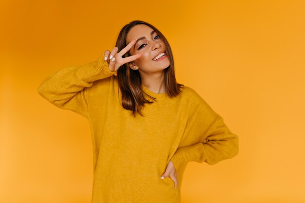 Alegre mujer bronceada posando con el signo de la paz. Fascinante chica caucásica en suéter amarillo sonriendo.