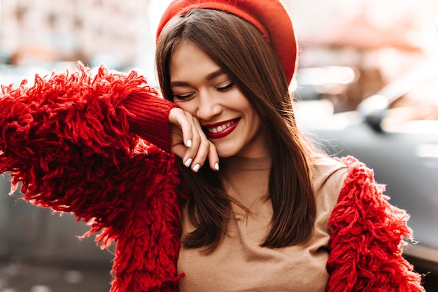 Alegre mujer de cabello oscuro con lápiz labial color burdeos vestida con elegante chaqueta de lana de gran tamaño y sombrero rojo se ríe, cubriéndose el rostro con la mano.