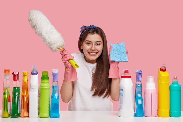 Alegre mujer de cabello oscuro lleva trapo y cepillo, sonríe alegremente, vestida con ropa casual, se sienta en un escritorio blanco con productos de limpieza