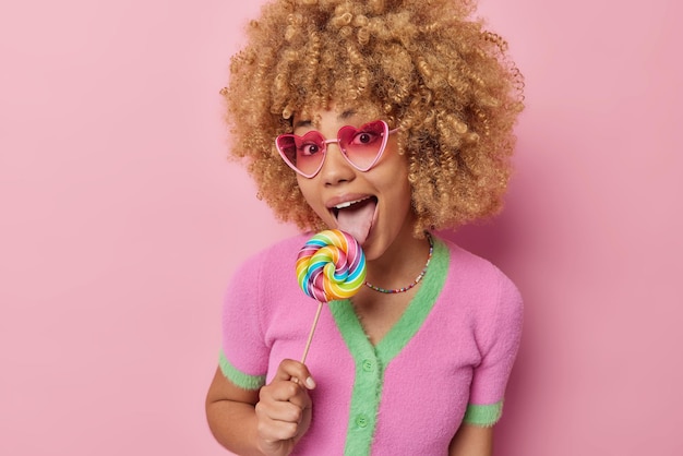 Foto gratuita alegre mujer de cabello rizado lame delicioso caramelo multicolor caramelo redondo siendo dulce adicto usa gafas de sol rosa de moda camisa casual aislada sobre fondo rosa concepto de nutrición de azúcar