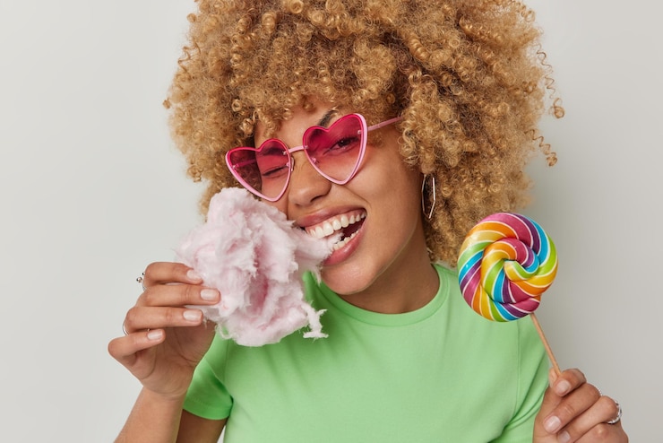 Foto gratuito alegre mujer de cabello rizado muerde delicioso algodón de azúcar con apetito sostiene dulces multicolores en un palo disfruta comiendo golosinas apetitosas usa gafas de sol en forma de corazón camiseta verde estilo de vida de verano