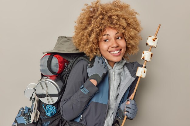 Alegre mujer de cabello rizado pasa tiempo libre en el campamento en caminatas por el bosque con mochila sostiene malvavisco asado en fogatas sonríe felizmente aislado sobre fondo gris Actividad de verano y estilo de vida