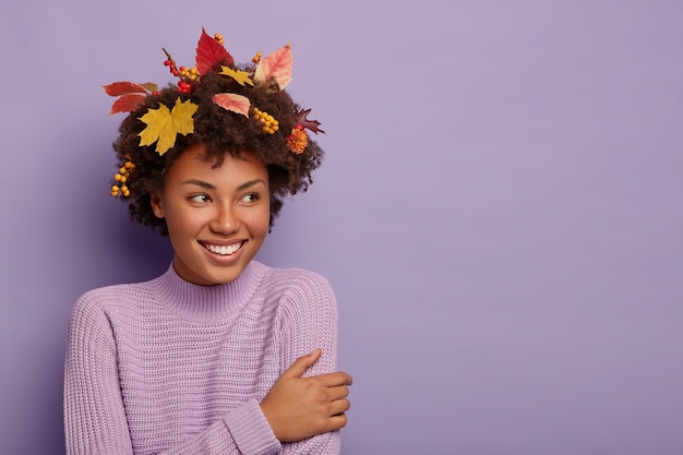 Alegre mujer de cabello rizado se vuelve a un lado, tiene una mirada alegre, está complacida con algo increíble, usa un atuendo casual, hojas de arce en la cabeza, sonríe agradablemente, posa en interiores