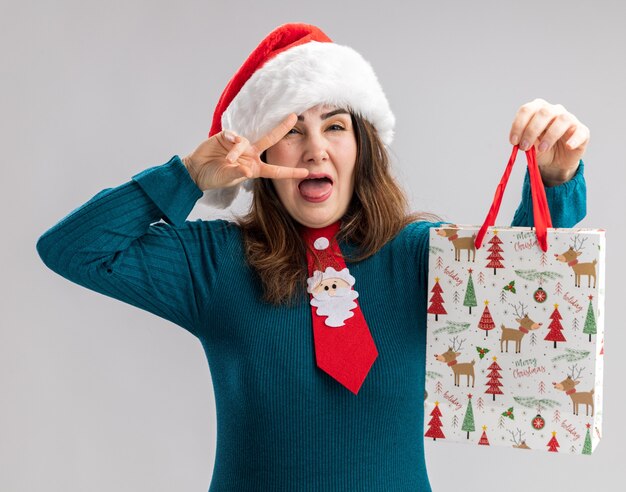 Alegre mujer caucásica adulta con gorro de Papá Noel y corbata de santa saca la lengua y sostiene una caja de regalo de papel aislada sobre fondo blanco con espacio de copia