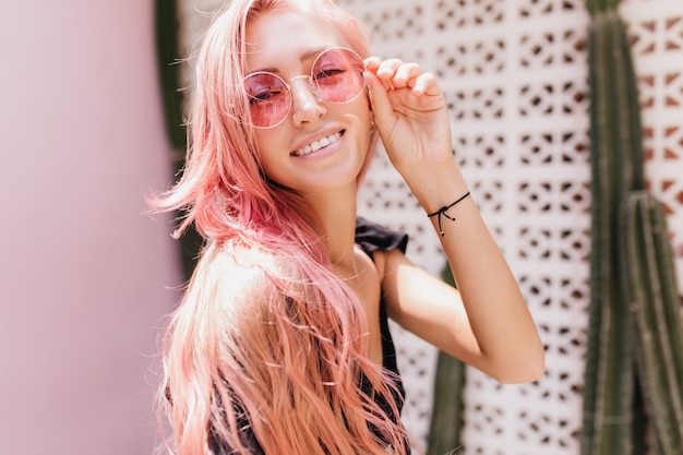 alegre mujer caucásica bronceada con cabello rosado. Dama europea complacida tocando sus gafas de sol mientras posa con plantas exóticas.